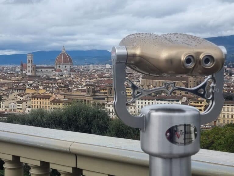 A public viewing telescope stands on a terrace overlooking Florence, with the Florence Cathedral dome and city rooftops visible in the background under a cloudy sky. The image emphasizes a panoramic viewpoint across the historic city.